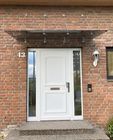 Modernes Glasvordach über einer weißen Eingangstür in einem Backsteingebäude, hergestellt von AP Glas GmbH aus Delbrück, für stilvolle und funktionale Hauseingänge.