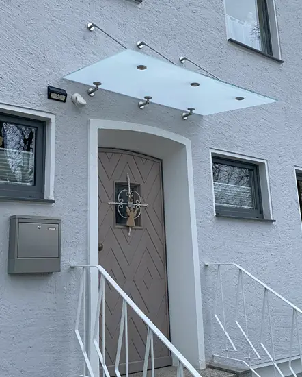 Glasvordach über der Eingangstür eines Hauses, montiert an einer weißen Wand, mit modernen Halterungen und einem klaren, eleganten Design.