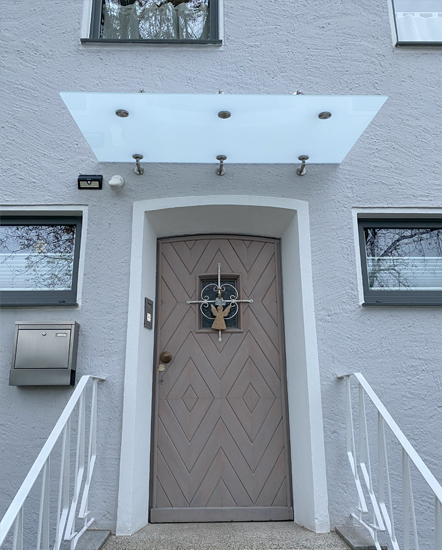 Moderne Glasvordach über der Eingangstür eines Hauses, montiert mit stilvollen Halterungen, sorgt für Schutz und ästhetischen Akzent. Ideal für zeitgemäße Architektur und hochwertige Eingangsbereiche.