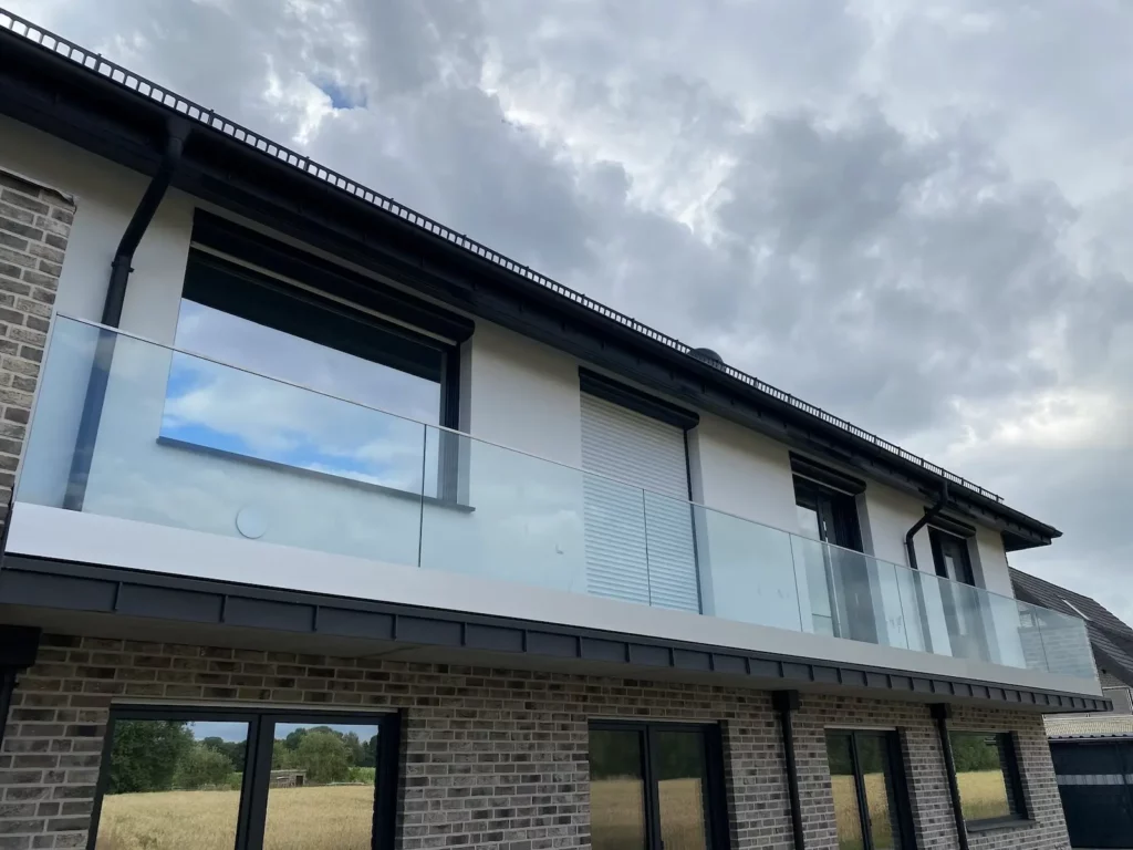 Moderne Glasbrüstung an einem Wohnhaus, die eine stilvolle und sichere Abgrenzung des Balkons bietet, umgeben von einem klaren Himmel und landwirtschaftlicher Landschaft im Hintergrund.